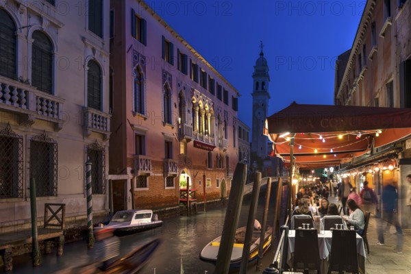 In the evening, lively restaurant on a canal with the tower of Santa Maria della Pietà in the back, Venice, Veneto, Italy