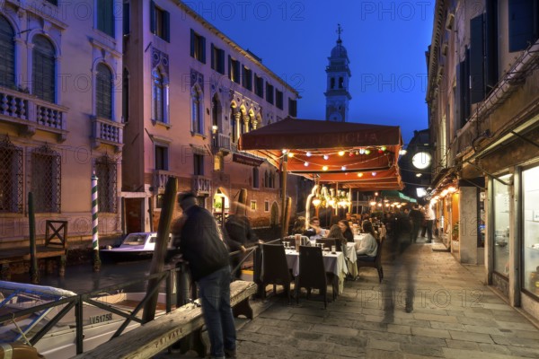 In the evening, lively restaurant on a canal with the tower of Santa Maria della Pietà in the back, Venice, Veneto, Italy