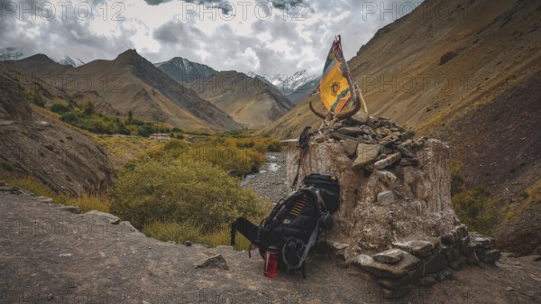Large hiking backpack leaning at a stone place of worship and Buddhist prayer flag on a rocky elevation with a mountain landscape in the background, trekking in Ladakh, Stok la, Himalayas, India