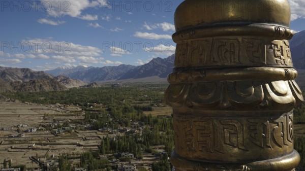 View from a monastery across the valley a golden prayer wheel in the foreground with a vast mountain landscape and clouds above, Leh, trekking in Ladakh, Himalayas, India