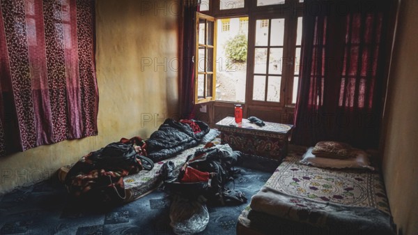 A simple room in a mountain village in the Indian Himalayas with two beds, backpack and cinder and a window with red curtains through which daylight falls, cozy colors and atmosphere, trekking in Ladakh, Stok la, Himalayas, India