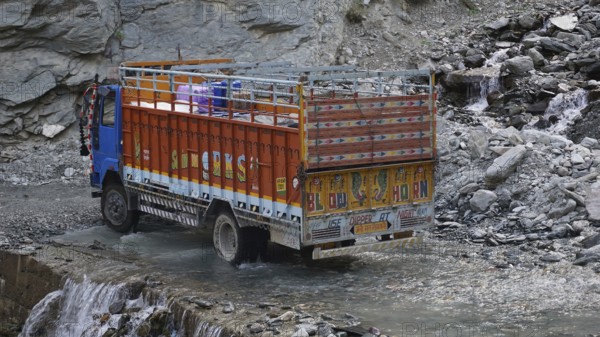 An Indian colorful truck crosses a shallow river on a road surrounded by rocky terrain on the Manali-Leh Highway, Leh, trekking in Ladakh, Himalayas, India