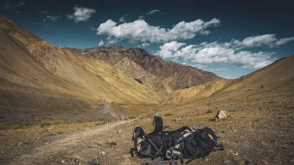 Hiking trail through a dry mountain landscape, a large backpack lying on the ground under a blue sky in front of a mountain panorama, trekking in Ladakh, Stok la, Himalayas, India