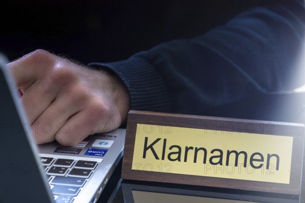 Clear name requirement Symbolic image: Brass sign with (digitally added) text KLARNAMEN next to a man sitting at a laptop