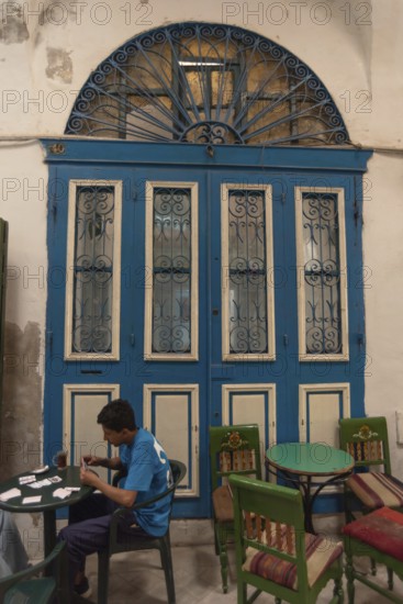 Tunis, Tunisia. 13th May 2024 A young Tunisian plays cards and drinks tea beside a traditional blue arched door in a café in the Medina of Tunis, Tunisia
