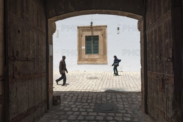 Tunisian men and women walk past some old wooden doors beside Bab Diwan the gate to Sfax Medina, Tunisia