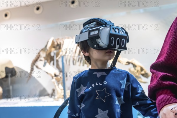 Young boy wearing a virtual reality headset, exploring an immersive experience at a museum with a dinosaur skeleton exhibit in the background, combining technology with education