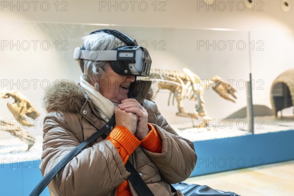 Senior woman wearing a vr headset, smiling with hands clasped while immersed in an interactive museum exhibit featuring dinosaur skeletons, blending technology and learning