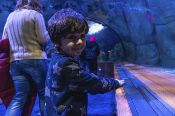 Young child with dark hair smiles, looking over shoulder while enjoying an immersive visit to an underwater tunnel in a large public aquarium, discovering marine life