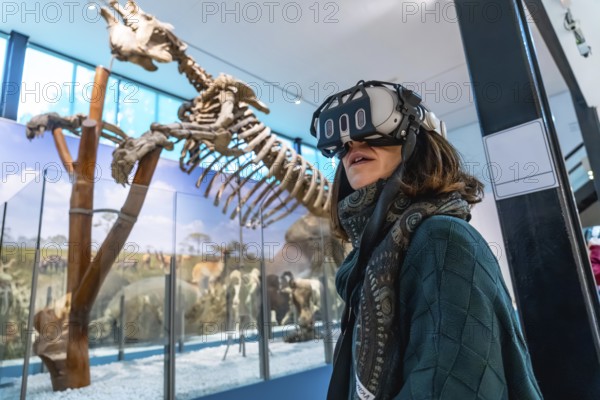 Woman wearing a virtual reality headset exploring an immersive exhibition with an ancient mammal skeleton and other animal displays in a modern museum environment