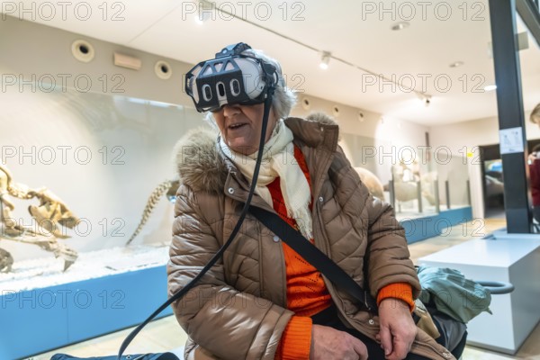 Senior woman wearing a vr headset, experiencing an immersive virtual reality exhibition in a modern museum with ancient animal skeletons in the background