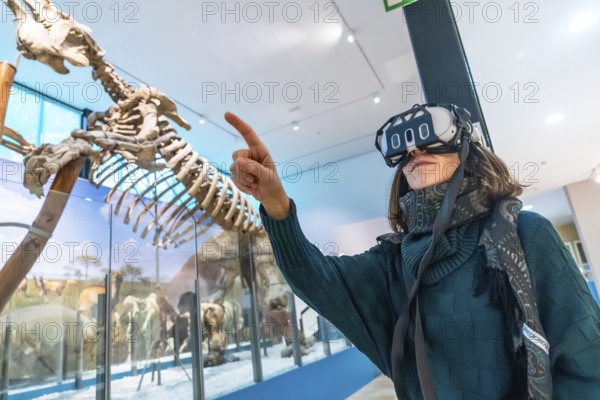 Woman wearing vr headset gestures and points at a life size dinosaur skeleton in a modern natural history museum, immersed in an educational mixed reality experience