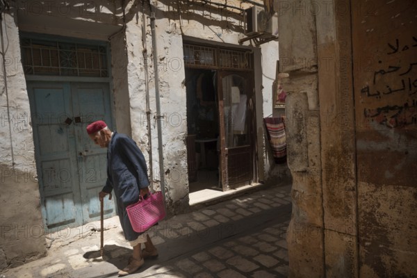 An old Tunisian man wearing a traditional red Chechia walks in the narrow alleyways of Sfax Medina, Tunisia, North Africa