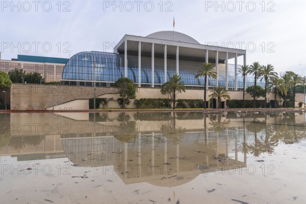 The palau de la musica music hall in valencia, spain, reflecting in a large permanent water feature or puddle with palm trees along the facade under a cloudy sky
