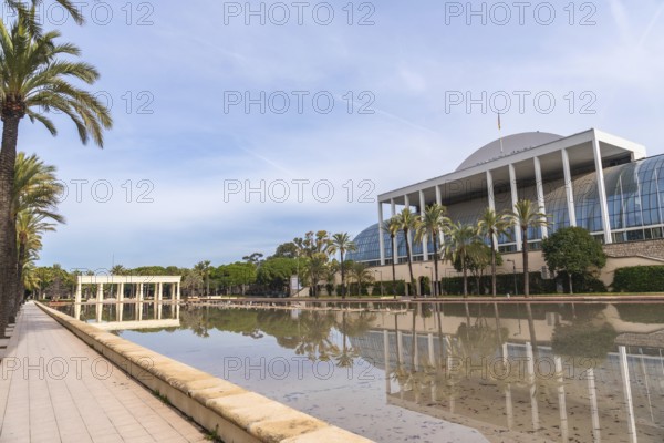 Palau de la musica building, an iconic landmark in valencia, reflecting in the turia gardens' calm waters surrounded by palm trees under a bright blue sky