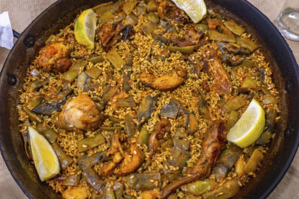 Traditional valencian paella cooking in a large pan with rice, chicken, rabbit, lima and green beans, saffron aroma and lemon wedges for a rustic mediterranean meal