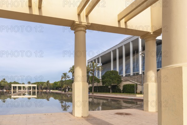 Palau de la musica building's modern architecture contrasts with classical columns of a pergola framing a pond reflecting green trees in the turia garden of valencia, spain