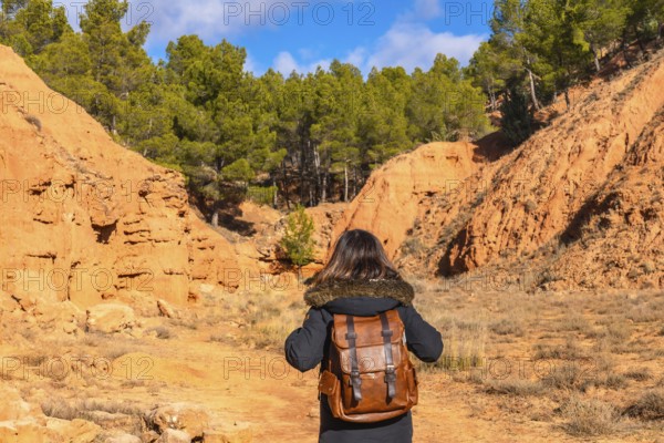 Hiker with a brown backpack walking through the unique red clay terrain of las arcillas natural park in teruel, spain, surrounded by eroding canyons and sparse green pine trees under a clear sky