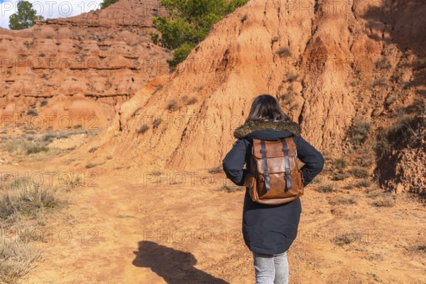 Woman with a backpack walking across a reddish clay landscape, discovering the unique geological formations and natural environment of the badlands in teruel, spain