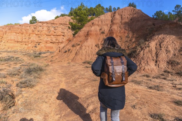 Woman with brown leather backpack and warm coat walking a dirt trail through las arcillas natural parks red clay badlands and pine trees under a clear blue sky