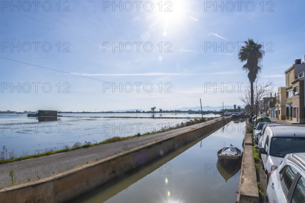 Traditional humble houses lining a narrow water canal with a small boat, reflecting the clear sky and bright sun, facing expansive flooded rice paddies in el palmar, albufera, valencia, spain