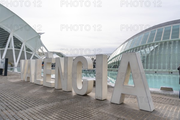 Oversized white valencia lettering on a modern promenade with the city of arts and sciences' futuristic architecture and reflective water features under a clear sky