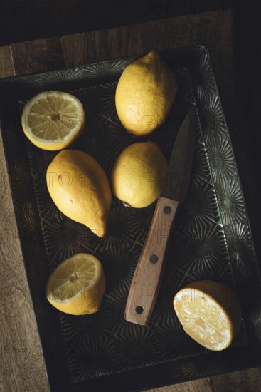 A rustic kitchen scene with fresh lemons on a vintage metal tray, accompanied by a wooden handled knife. The soft lighting enhances the textures of the lemons