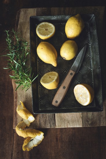 A rustic still life featuring fresh lemons on a vintage tray with a wooden handled knife and a sprig of rosemary. Lemon peels add texture, creating a warm, inviting scene