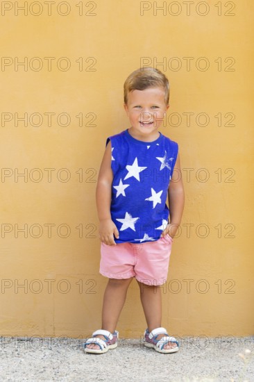 A cheerful young boy in a blue shirt with white stars poses against a vibrant yellow wall His pink shorts and sandals add a playful touch to the summery scene