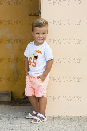 A young boy wearing a white t-shirt with a toucan design and peach shorts leans against a wall He smiles brightly, exuding joy and carefree summer vibes