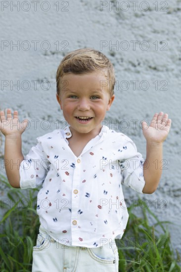 A cheerful child with an adorable smile raises their hands playfully against a textured wall backdrop, wearing a patterned shirt and jeans, enjoying a sunny day outdoors