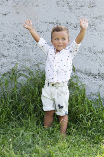 A cheerful young child stands with arms raised in a grassy garden Dressed in shorts and a patterned shirt, the child exudes joy and playfulness against a textured wall backdrop