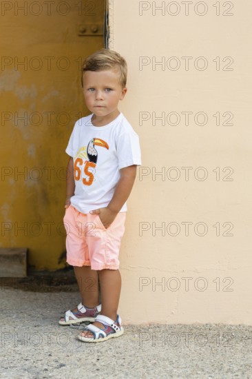 A young boy stands thoughtfully against a pastel wall, wearing a white toucan t-shirt, pastel shorts, and sandals His casual summer outfit creates a relaxed mood