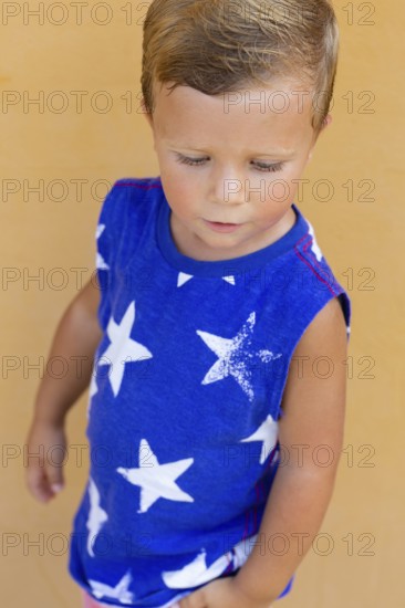 A young child wearing a blue shirt with white star patterns stands against a light orange wall The child looks down, capturing a moment of thoughtfulness