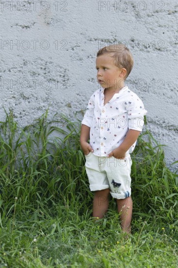 A young boy stands against a gray concrete wall, dressed in a white shirt and shorts The grass at his feet adds a natural touch His pose is relaxed and thoughtful