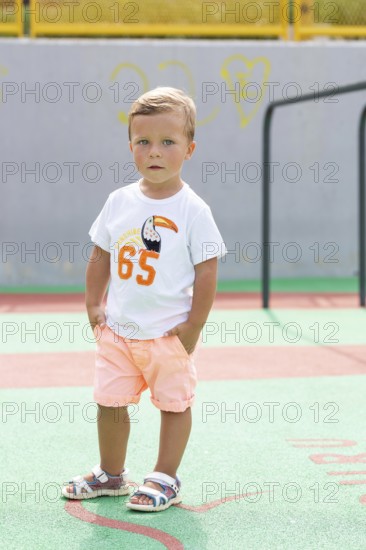 A young boy stands confidently on a vibrant playground, wearing a white T-shirt and peach shorts The sunny day highlights his playful sandals and summer spirit