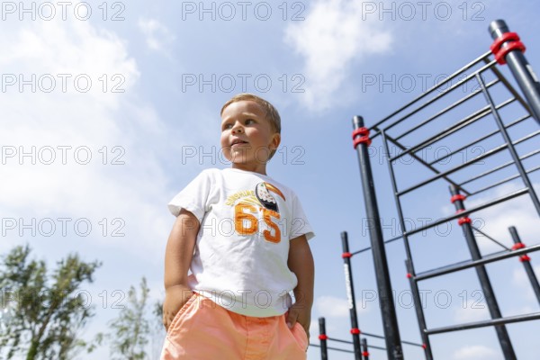 A young child stands confidently in a playground, wearing a colorful outfit against a clear blue sky with clouds The child is near climbing bars, indicating play and adventure