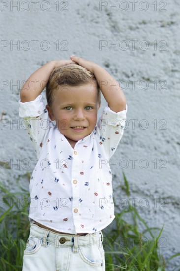 A cheerful child stands against a textured wall, hands on head, wearing a white shirt with small patterns The casual and natural setting highlights the playful spirit