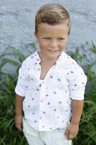 A cheerful young boy with a charming smile stands against a gray wall, dressed in a playful patterned white shirt Green grass complements the background