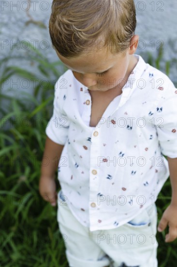 A young boy wearing a white shirt with colorful prints stands amidst green foliage His gaze is downward, creating a serene and candid outdoor moment