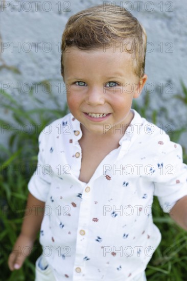A cheerful young boy in a patterned white shirt smiles brightly at the camera while standing outside The green grass and textured wall in the background add freshness to the scene