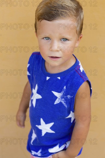 A young boy with blue eyes gazes upwards, wearing a blue shirt with white star patterns His expression is curious and innocent, against a warm beige background, creating a joyful image