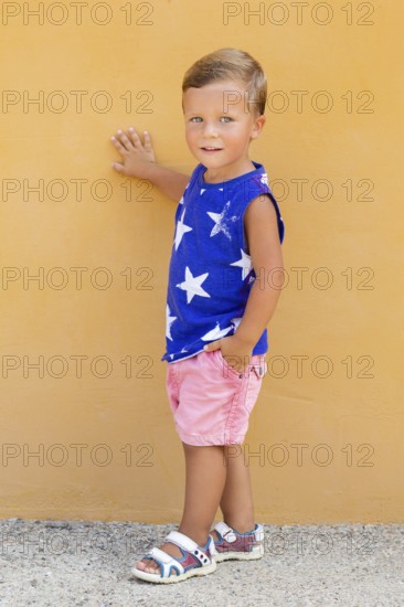 A young child stands against a vibrant orange wall, wearing a blue sleeveless shirt with white stars, pink shorts, and sandals, projecting a confident and playful vibe