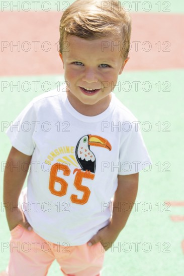 A cheerful child wearing a white T-shirt with a toucan design and number, standing outdoors on a vibrant background The sunlight highlights the kid's playful smile