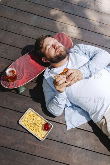 A man lies on wooden decking, enjoying a burger with a tray of fries and a drink resting on a skateboard beside him. The scene captures a relaxed, outdoor barbecue vibe