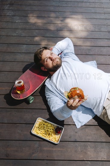 A person enjoying a relaxing moment on a wooden deck, holding a burger. Nearby, there's a skateboard, fries, and a drink, capturing the essence of a laid-back barbecue day