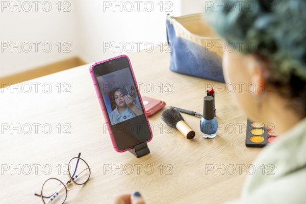 A transgender man records a makeup tutorial with his mobile phone, showcasing various cosmetic products laid out on a table, including brushes, lipstick, and eyeshadow, to guide viewers through his makeup application process