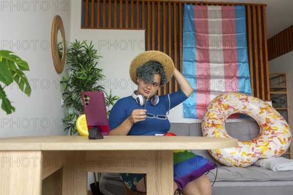Transgender man hosting a virtual meeting from home, displayed on a smartphone. He is sitting at a wooden table, wearing a straw hat and a blue shirt. A pride flag is draped as a backdrop, symbolizing LGBTQ+ support