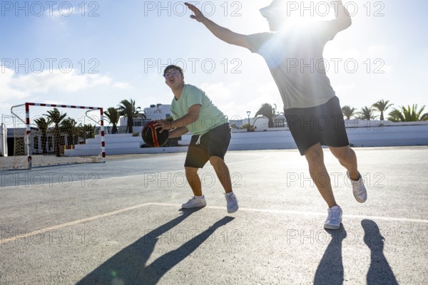 Down syndrome teenager joyfully plays basketball with his brother on an outdoor court The sun shines brightly, creating a warm and supportive environment