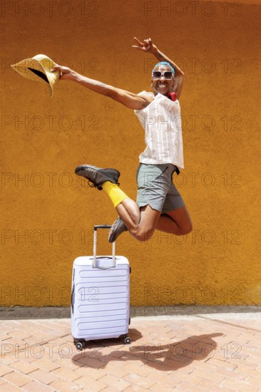 A joyful LGBTQIA+ man jumps exuberantly with a suitcase, ready to embark on a vibrant holiday trip. Bright attire and mural create a lively, celebratory mood
